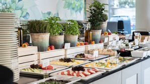 Un bufé con diversas opciones de comida, como ensaladas, carnes y frutas, dispuesto en un mostrador decorado con plantas y vistas al exterior.