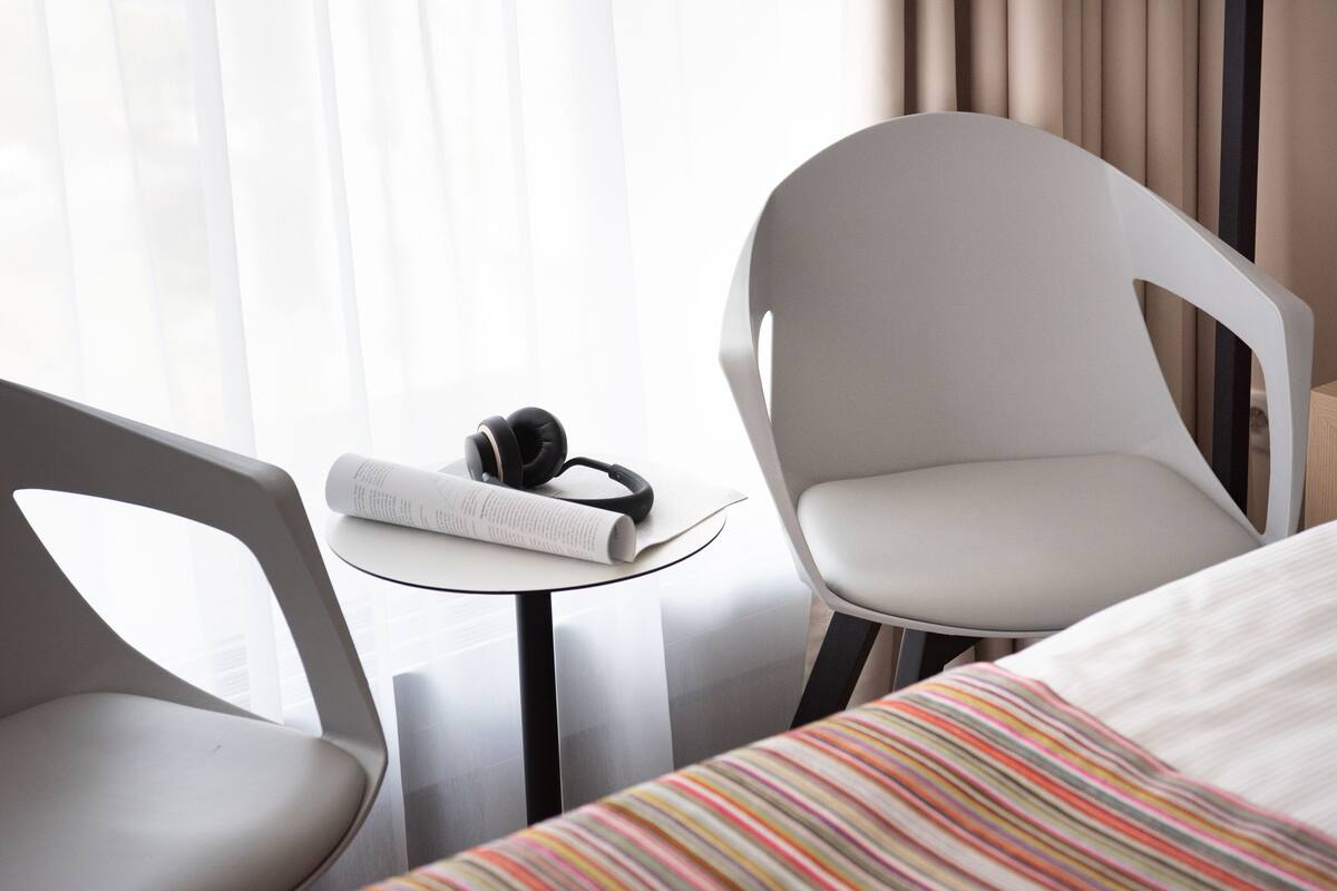 Une chambre d’hôtel moderne, équipée d’un lit, de deux fauteuils, d’une petite table avec un casque audio et un livre, ainsi que d’une fenêtre avec des rideaux.