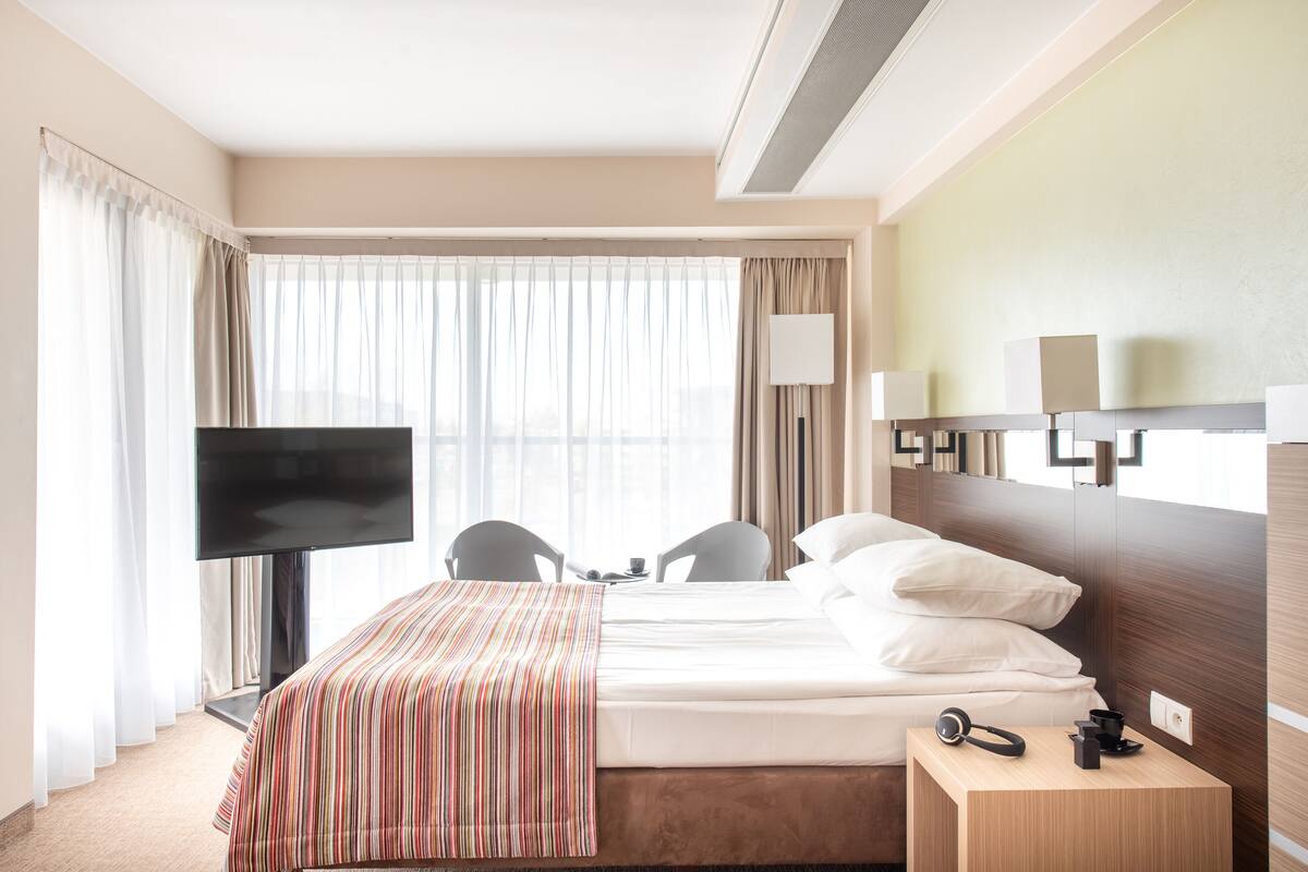 Une chambre d’hôtel avec un lit, une télévision, un bureau avec une chaise, une lampe et une fenêtre avec des rideaux.