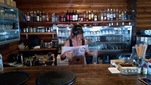 Een bar met drankflessen op planken, een gekoeld display en een toonbank waar iemand een drankje aan het maken is.