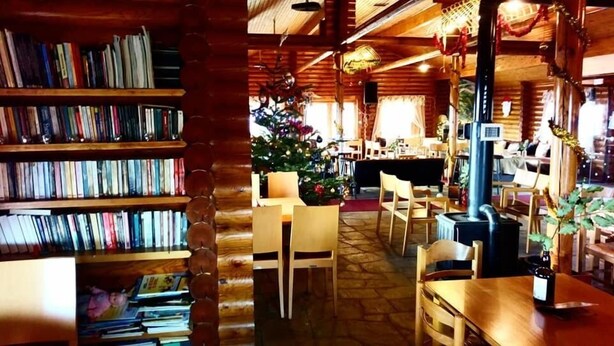 Een gezellig interieur met een houten boekenkast, een kerstboom en een eethoek met stoelen en tafels.