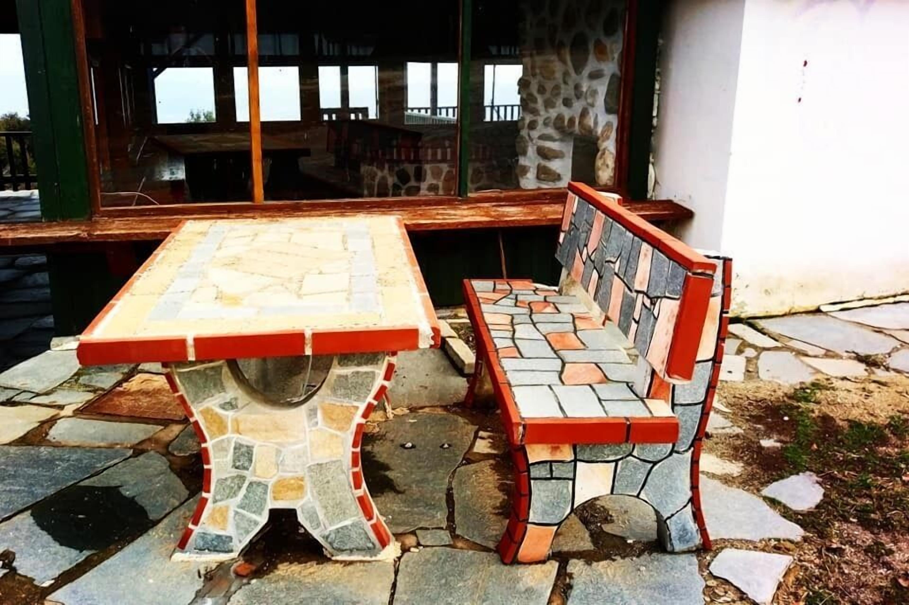 Eine Steinsitzbank und ein Tisch mit Mosaikmuster, draußen aufgestellt, mit einer Steinmauer und Fenstern im Hintergrund.