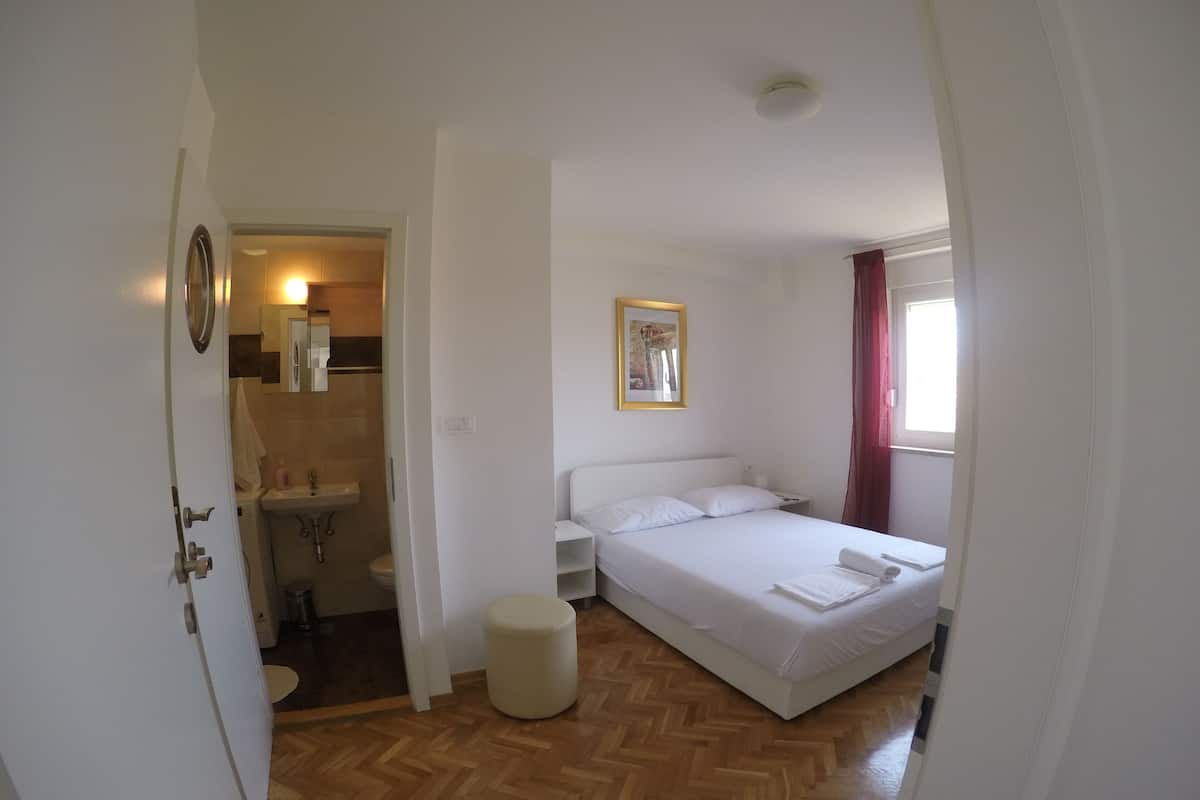 A bedroom with a bed, a bedside table, a nightstand, a round ottoman, a window with curtains, and a bathroom visible through an open door.