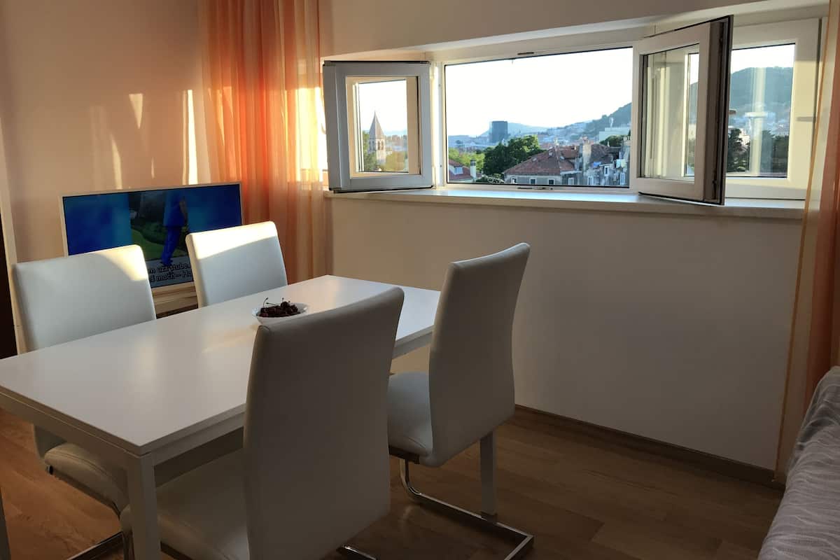 A modern room with a dining table, white chairs, a TV, and a window with a city view.