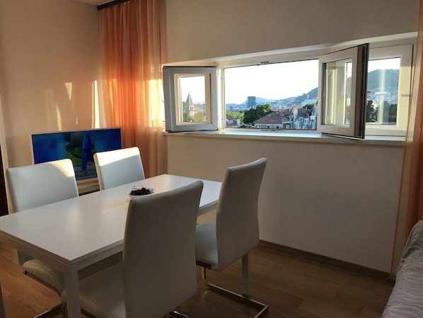 A modern room with a dining table, white chairs, a TV, and a window with a city view.