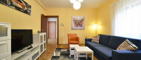 Una sala de estar con televisor, sofá y sillones, un cuadro en la pared y una puerta que da a otra habitación.