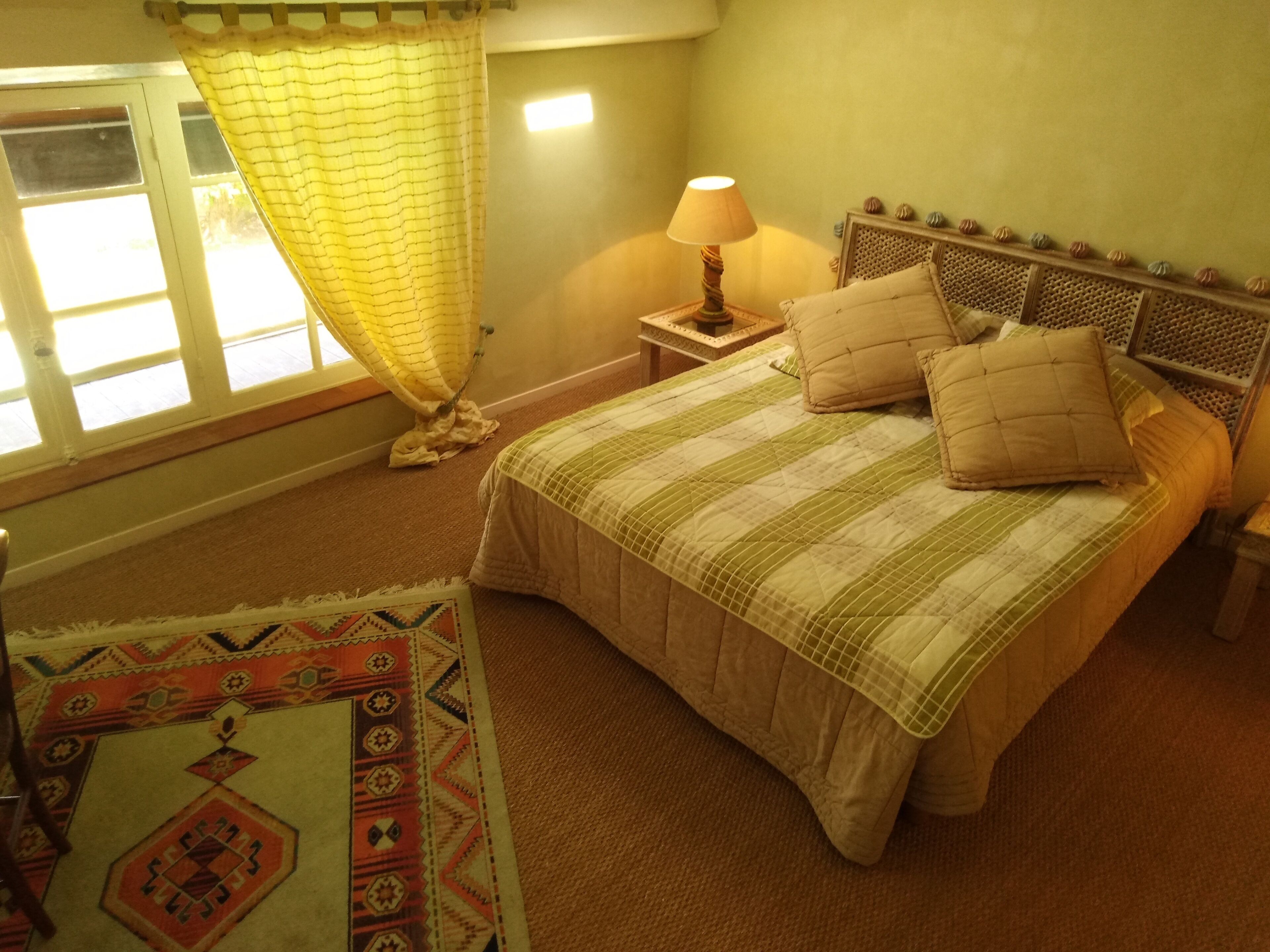 A bedroom with a bed, a nightstand, a lamp, a window with curtains, and a patterned rug.