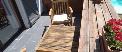Une terrasse en bois avec une table et deux chaises, entourée de plantes en pot et d’une piscine.