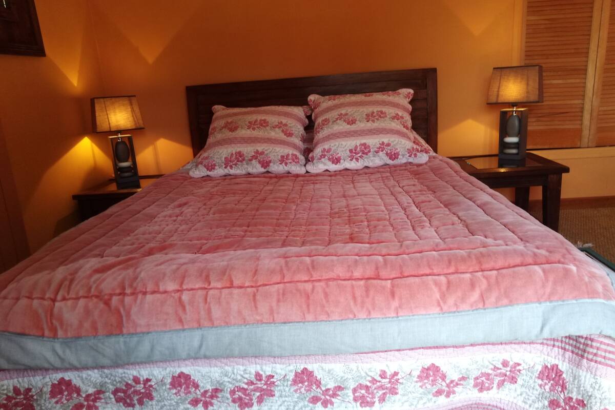 En säng med ett rosa täcke och blommig dekorerade kuddar, två sänglampor och en sänggavel i trä.
