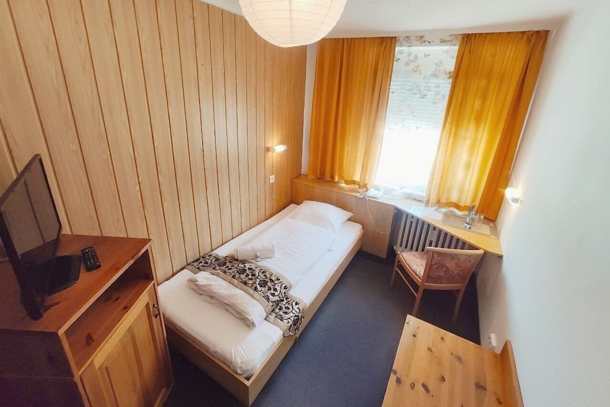 Ein Einzelbett mit Holz-Kopfteil, ein Schreibtisch mit Stuhl, ein Fernseher und ein Fenster mit Vorhängen.