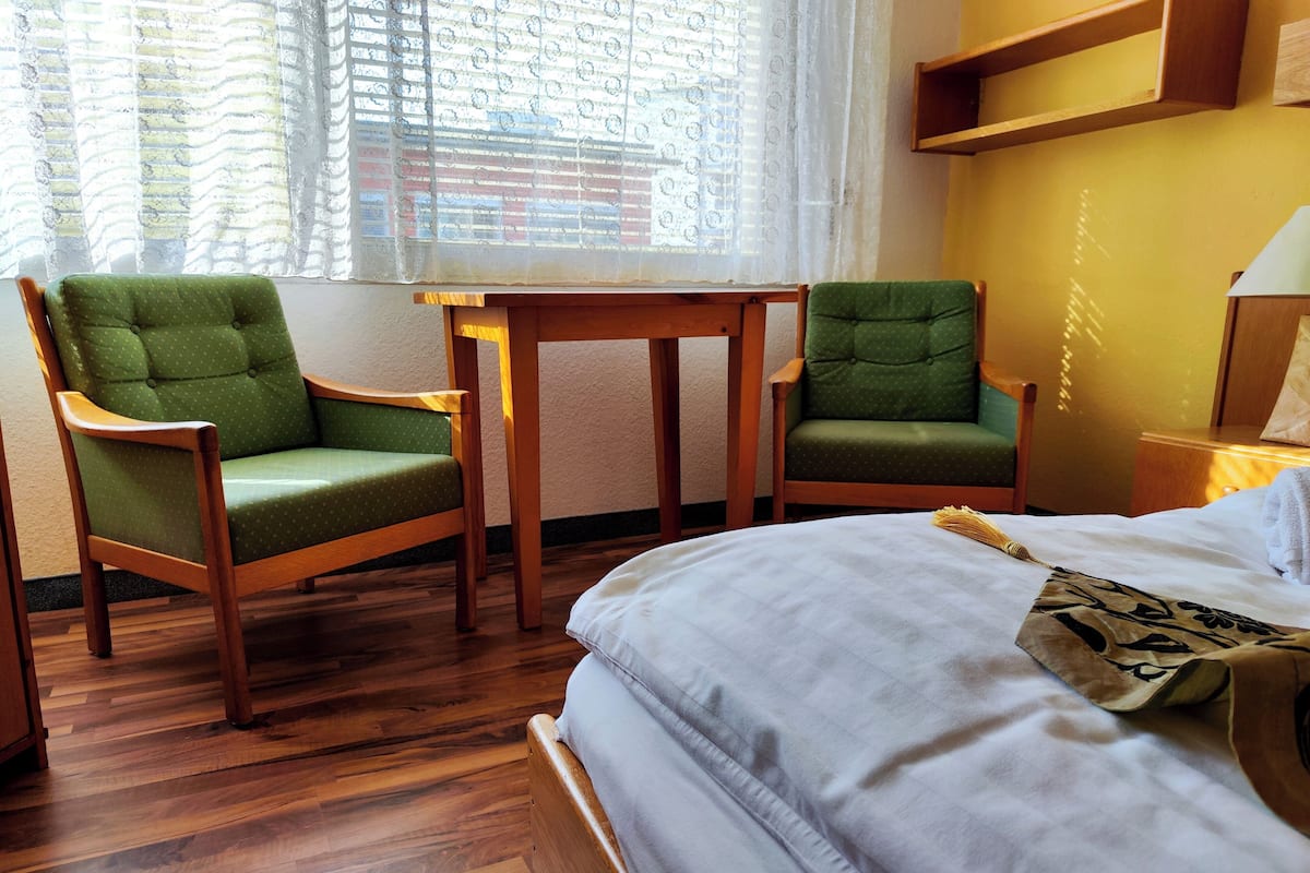 Ein Hotelzimmer mit einem Bett, zwei grünen Polsterstühlen, einem kleinen Holztisch und einem Fenster mit gemusterten Vorhängen.
