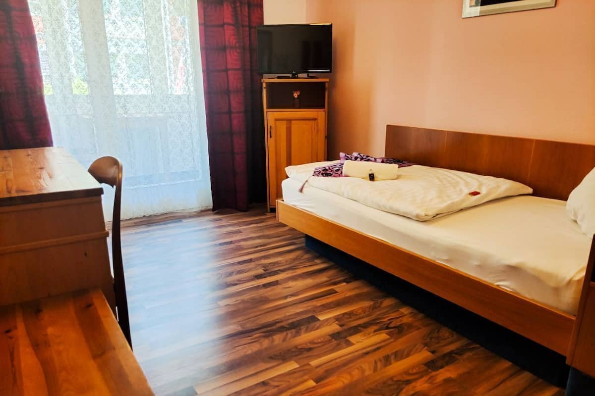 Ein Zimmer mit Holzboden, einem Bett mit weißer Bettwäsche, einem Holzkasten, einem Fernseher darauf und einem Sessel.