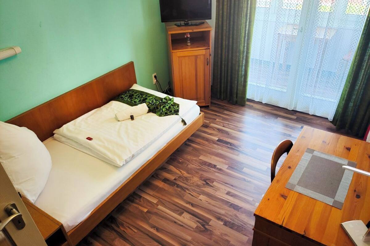 Ein Zimmer mit einem Holzbett, einem Schreibtisch aus Holz, einem Fernseher auf einem Schrank und Blick ins Freie durch ein Fenster mit Vorhängen.