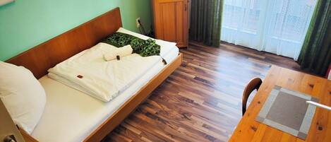 Una habitación con una cama de madera, un escritorio de madera, un televisor sobre un mueble, y vistas al exterior a través de una ventana con cortinas.