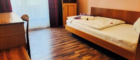 A room with a wooden floor, a bed with white bedding, a wooden cabinet, a television on top of the cabinet, and a chair.