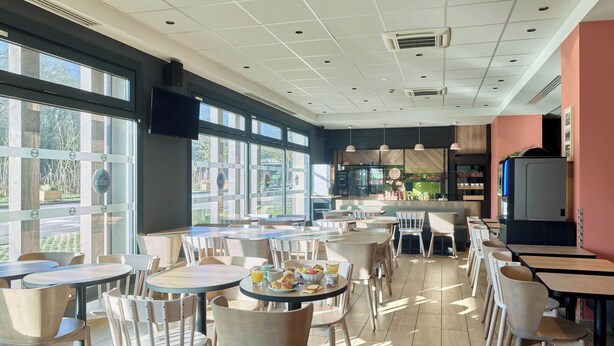 A restaurant with round tables and chairs, large windows, and a kitchen area. - B&B HOTEL Paris Meudon Vélizy (Meudon)