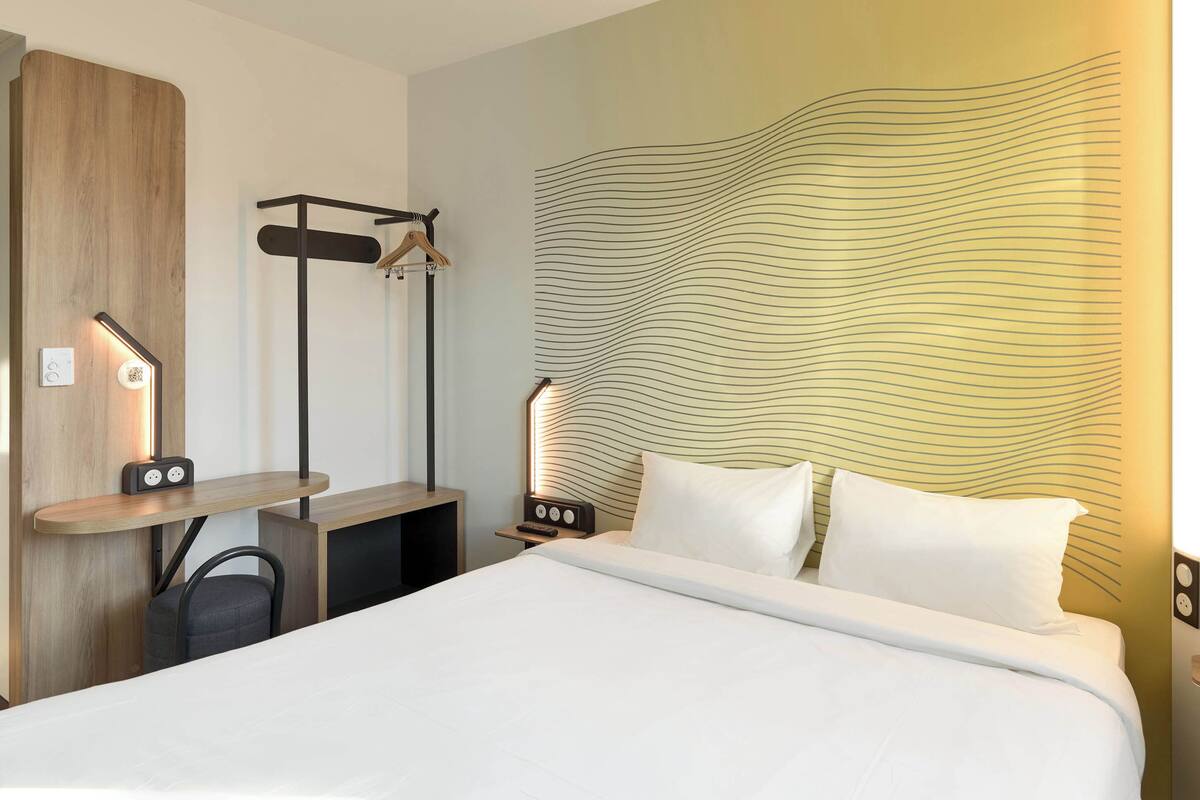 Habitación de hotel con una cama grande, un escritorio de madera, una silla, una mesita de noche con lámpara y una pared con diseño ondulado.