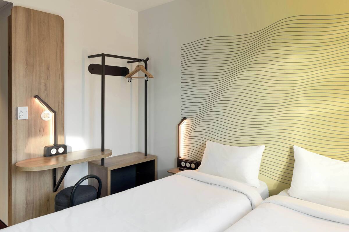 Habitación de hotel con cama, mesita de noche y escritorio con silla. Se observa un perchero y una pared con diseño ondulado.