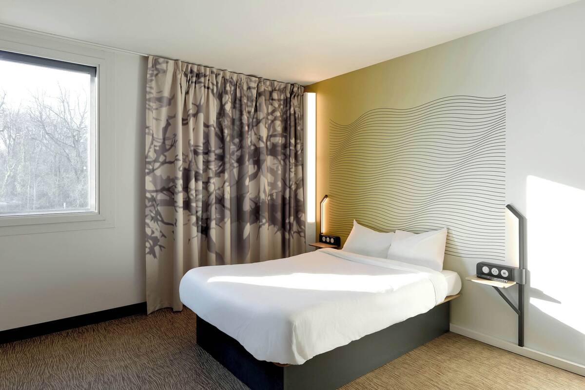 Habitación de hotel moderna con una cama grande, una mesita de noche con lámpara y vistas a árboles por la ventana.