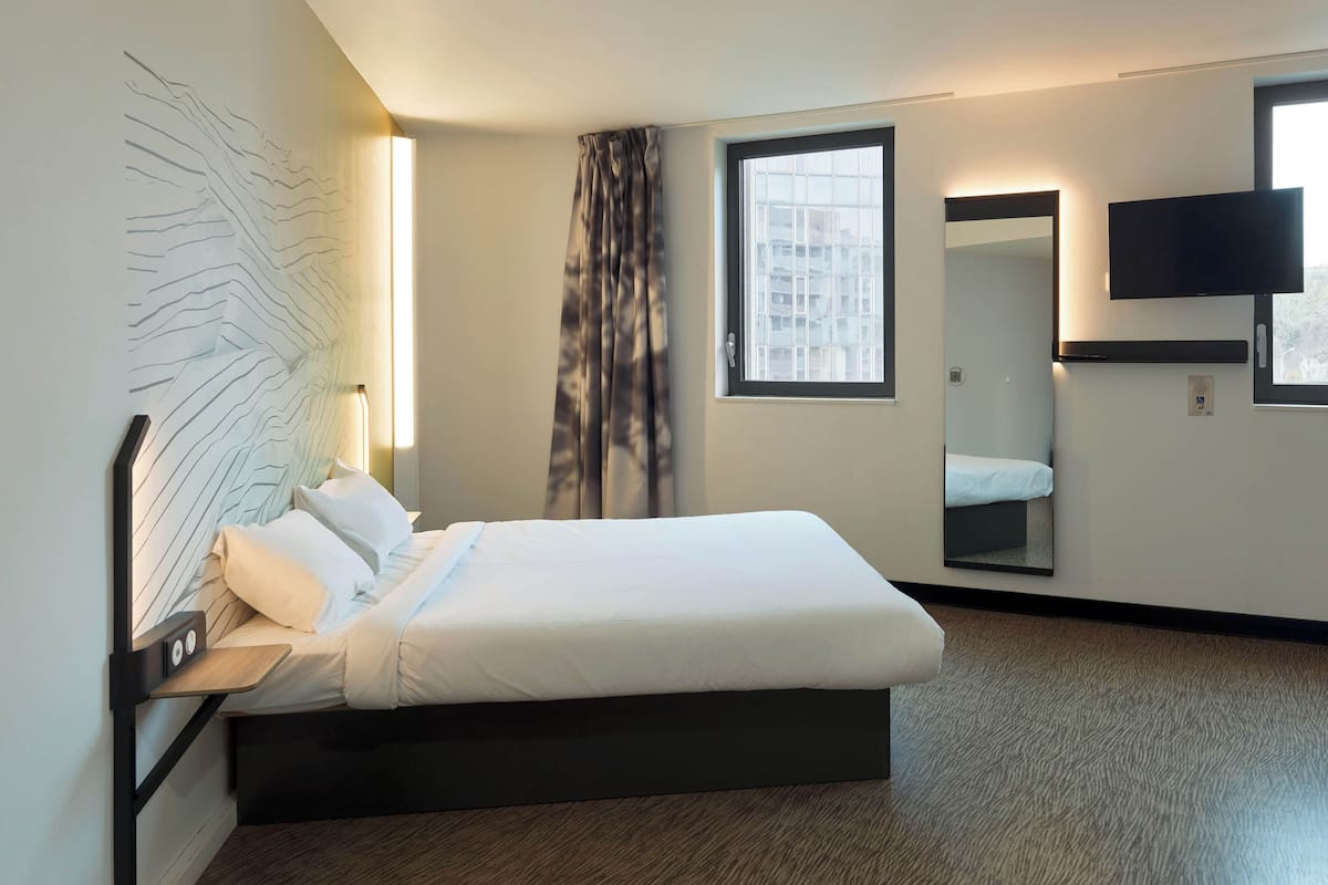 A hotel room with a bed, a bedside table, a TV mounted on the wall, a window, and a mirror.