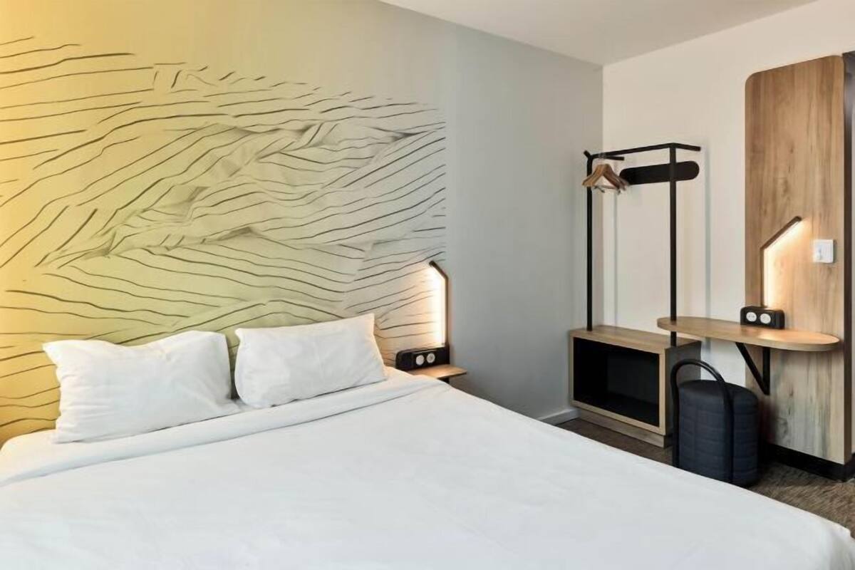 A modern hotel room with a large bed, a headboard with a unique design, bedside tables with lamps, a wardrobe, and a luggage rack.