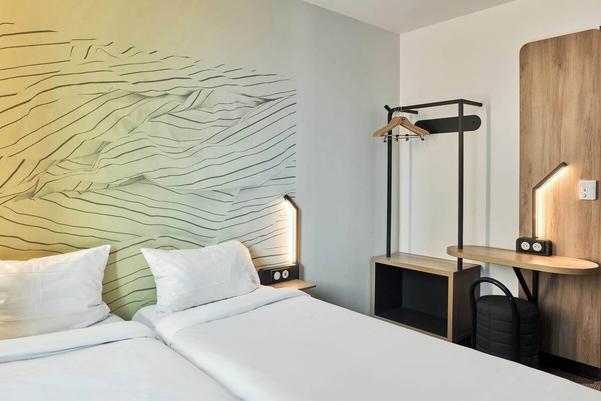 A modern hotel room with a bed, bedside tables, a wooden wardrobe, and a hanging rack.