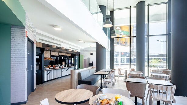 Una cafetería moderna con mesas y sillas redondas, una barra con comida y bebidas, y amplios ventanales con vistas a la ciudad.
