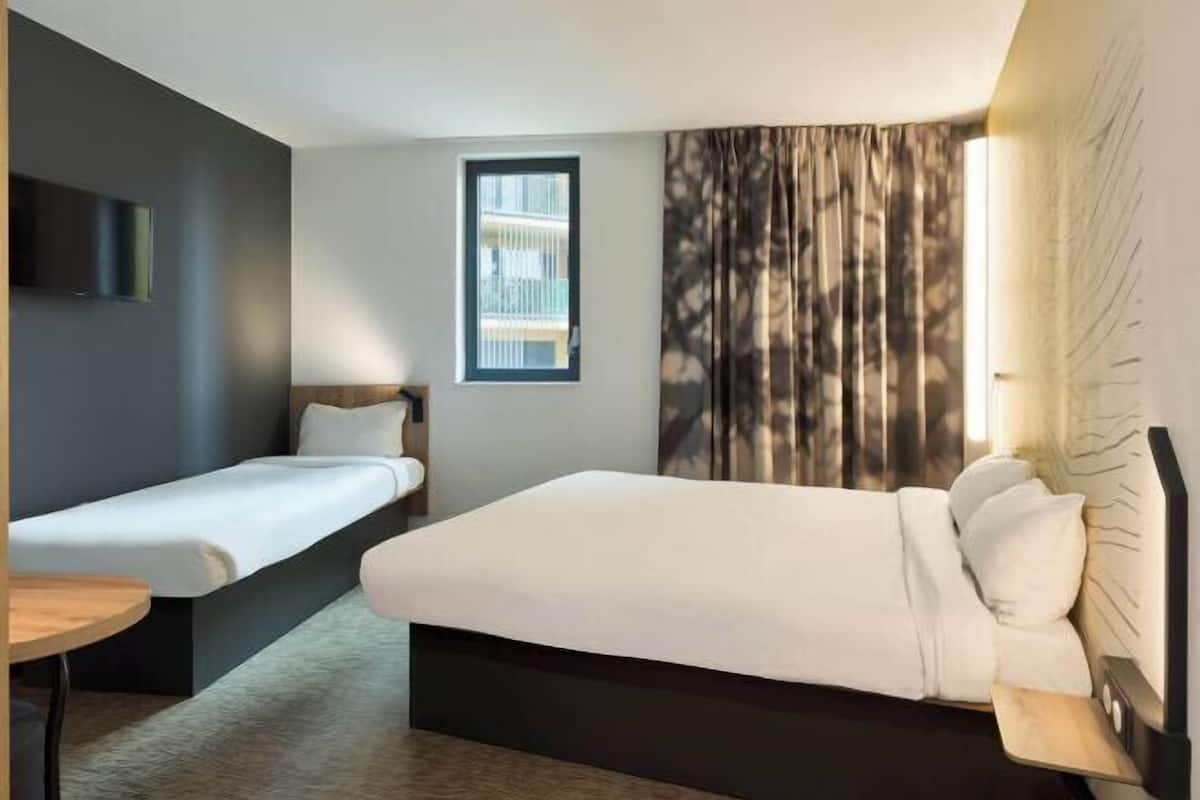 A hotel room with a single bed, a smaller bed, a desk, a TV, and a window with curtains.