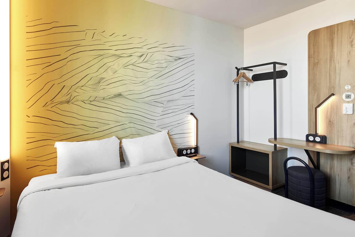 Habitación de hotel con cama, una obra de arte en la pared, una mesita de noche de madera, una lámpara y un perchero.