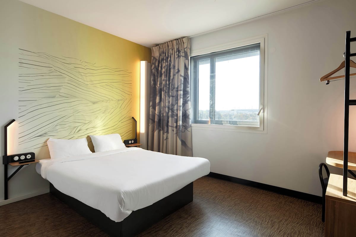 Una habitación de hotel con una cama grande, una mesita de noche con lámpara, una ventana con cortinas y vistas a la ciudad.