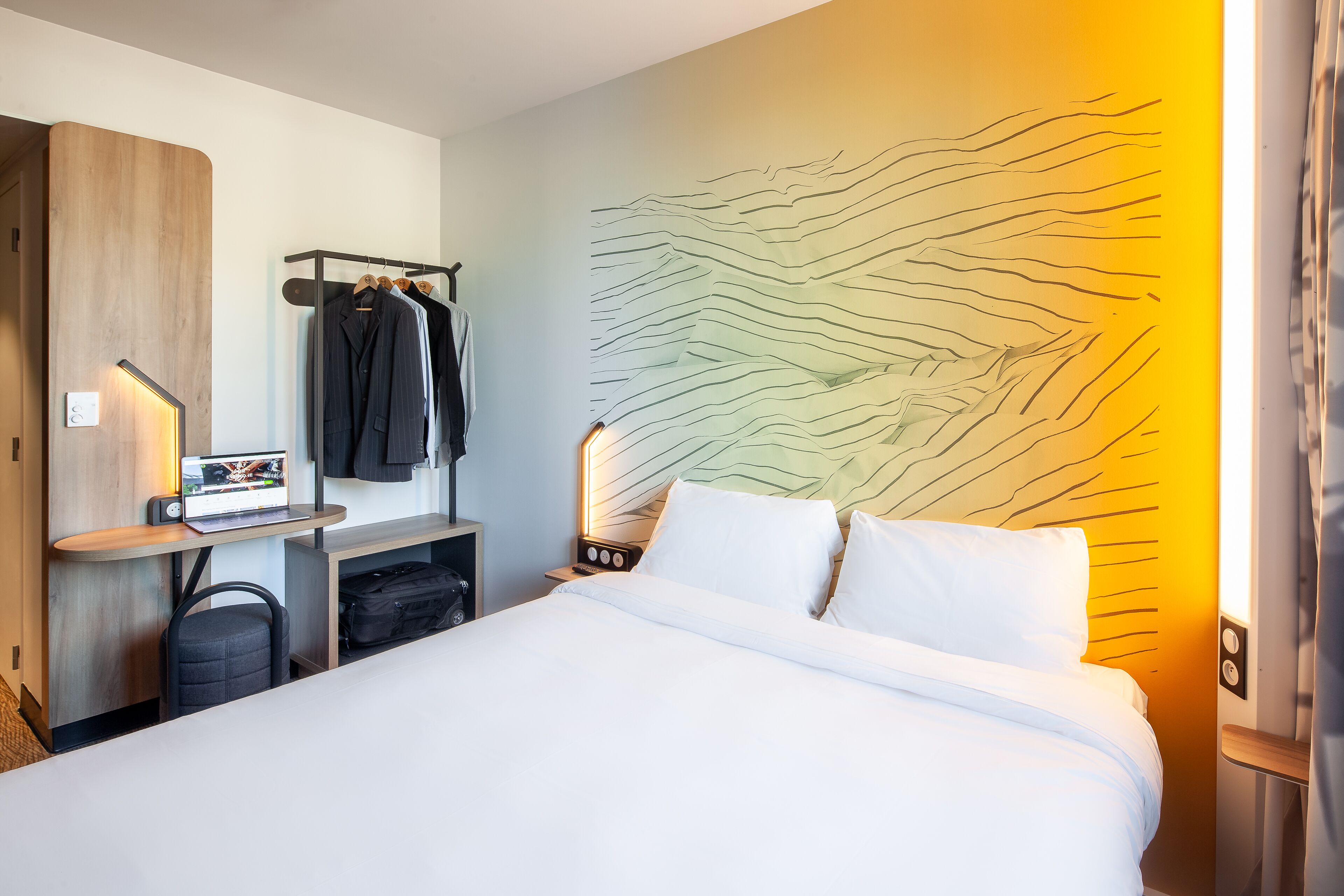 A hotel room with a bed, a desk with a laptop, a coat rack, and a wall with a textured design.