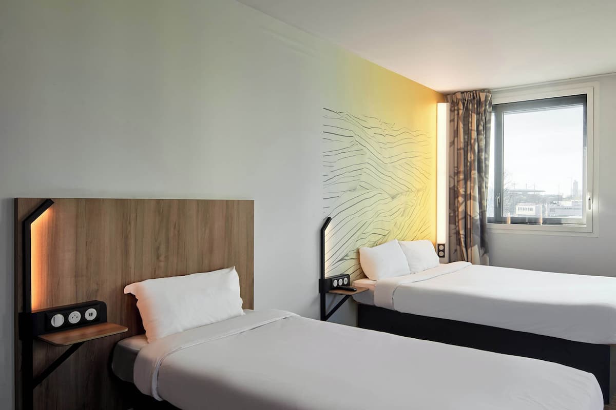 Habitación de hotel con dos camas individuales, cabecero de madera, una obra de arte en la pared, una ventana con cortinas y una mesita de noche con toma de corriente.