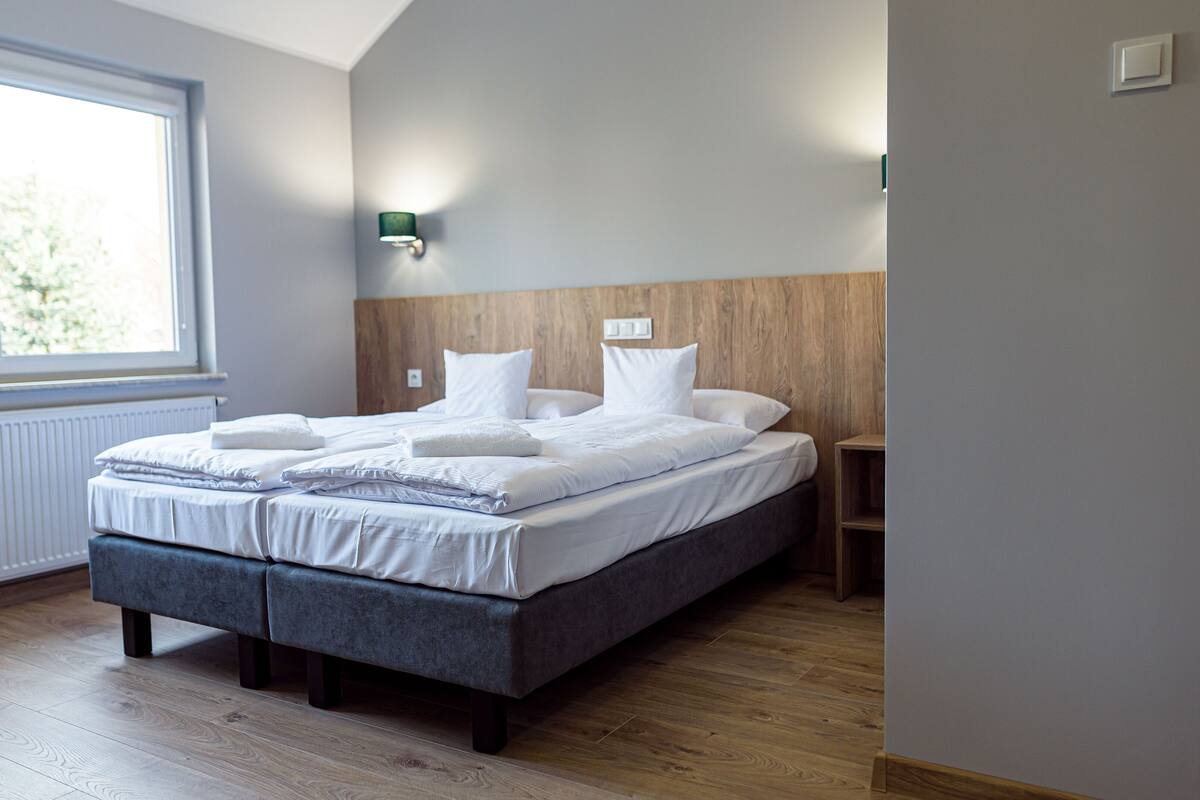 A neatly made bed with white linens and pillows, a wooden headboard, a bedside table, and a window with a view of greenery.