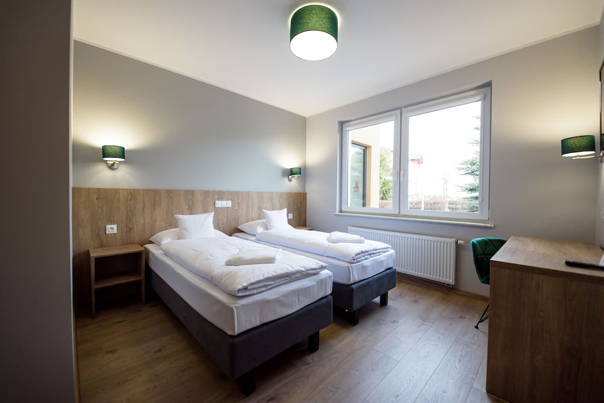 A modern hotel room with two beds, wooden headboards, a window with a view, and a small desk with a chair.