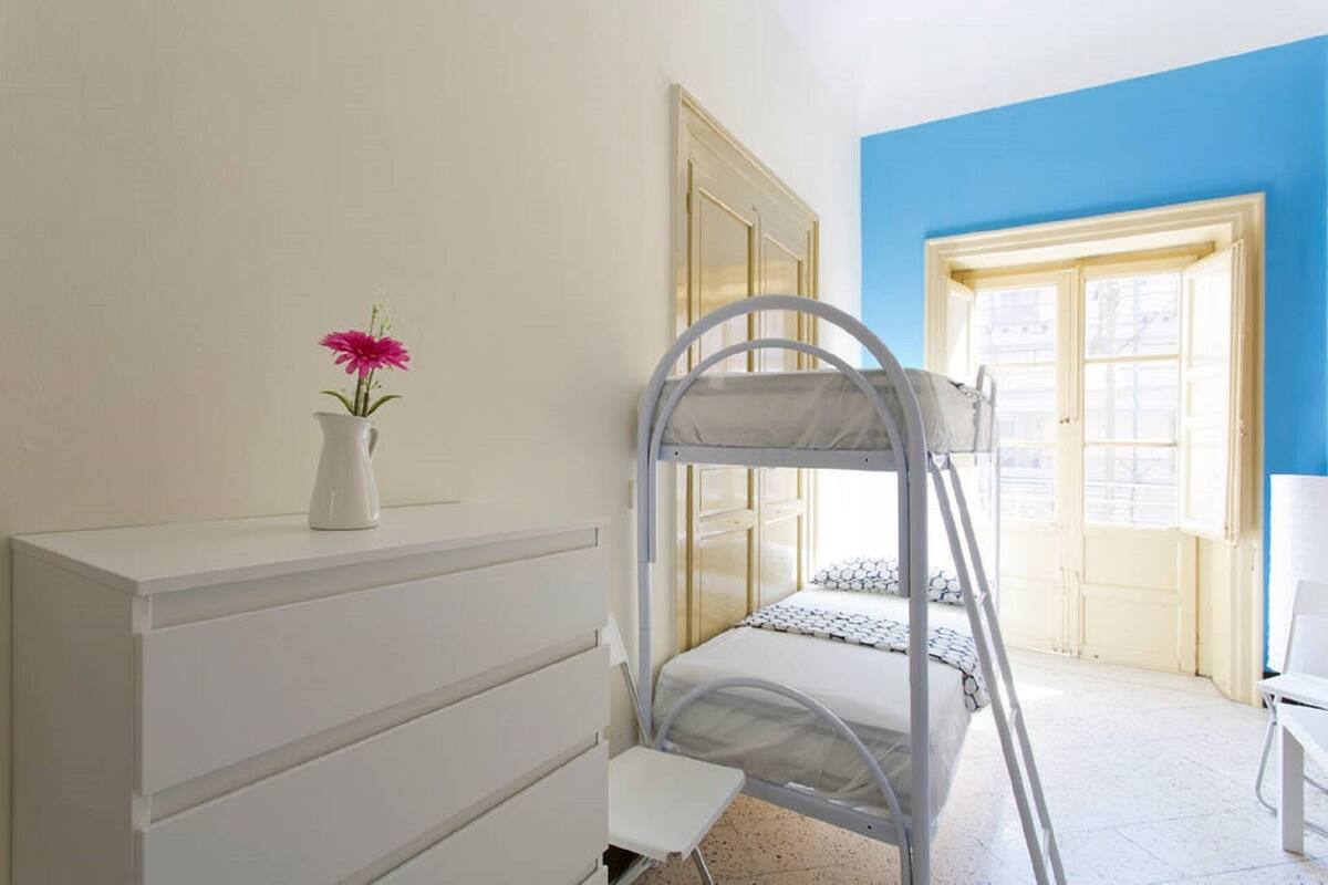 Une chambre avec un lit superposé, une commode blanche, un vase avec une fleur et une fenêtre donnant sur un immeuble.