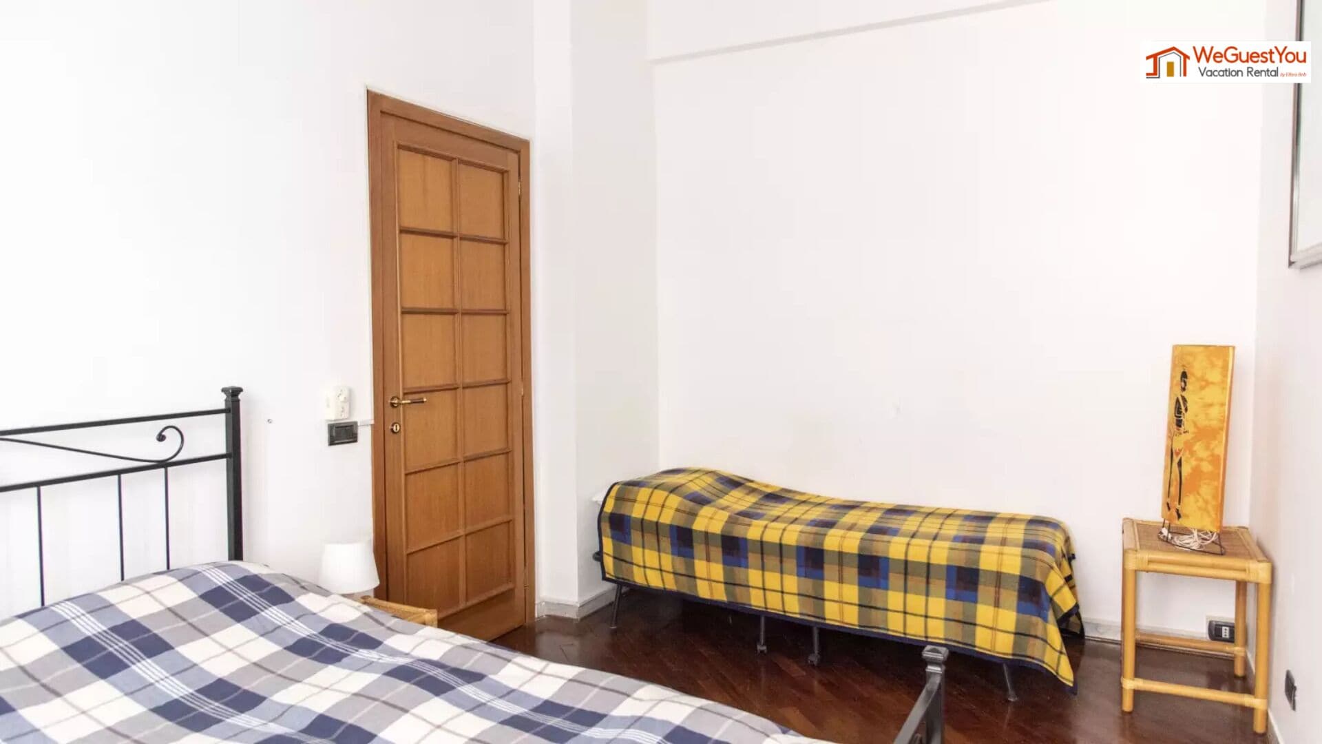 Une pièce comprenant un lit, une porte en bois, une petite table avec une lampe et une œuvre d’art accrochée au mur.