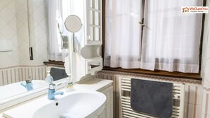 A bathroom with a white sink, a mirror, a towel rack, and a window with sheer curtains. - Flavia Home - 3 bedroom in Flavia Home Holiday (Rome)
