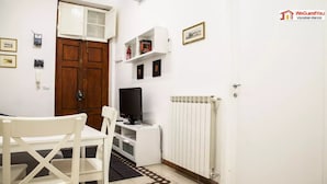 Una habitación pequeña y limpia con una puerta de madera, paredes blancas, un radiador blanco, un escritorio con una computadora y una silla.