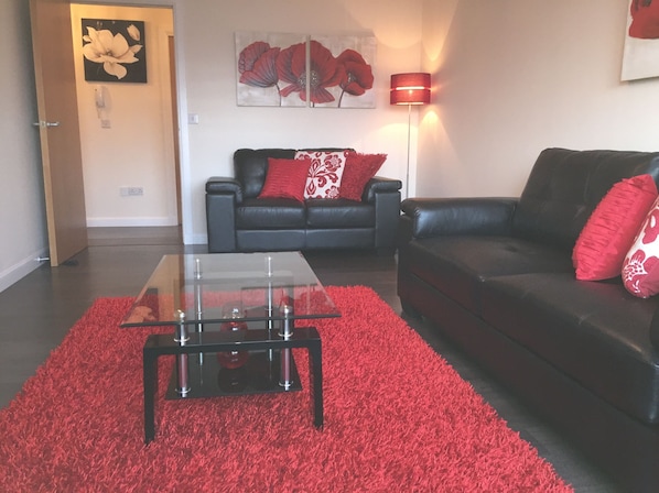 A living room with a glass coffee table, a red rug, a black sofa with red cushions, and a floor lamp.