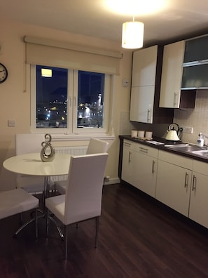 A modern kitchen with white cabinets, a dining table with chairs, and a view of the city at night through a window.