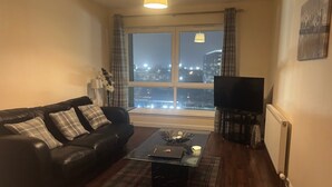 Uma sala de estar com um sofá de couro preto, uma mesa de centro de vidro e uma televisão. Há uma janela com vista para o horizonte urbano à noite.