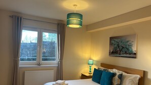 Quarto de hotel com cama, janela com cortinas, radiador, mesa de cabeceira com abajur e um quadro na parede.