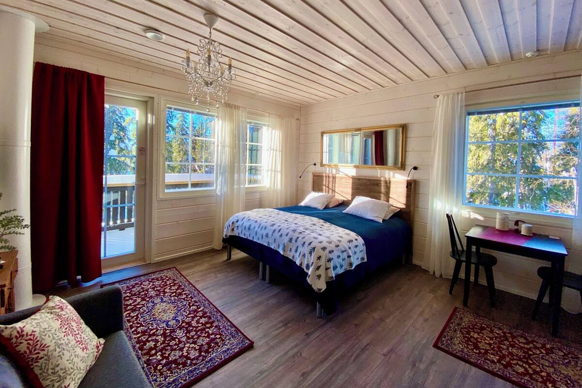 A bedroom with a wooden ceiling, a bed with a blue and white bedspread, a wooden headboard, a desk with a chair, and large windows with curtains.