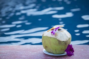 A green coconut with a straw and purple flower on a white plate, placed on a red surface with a blue water background.