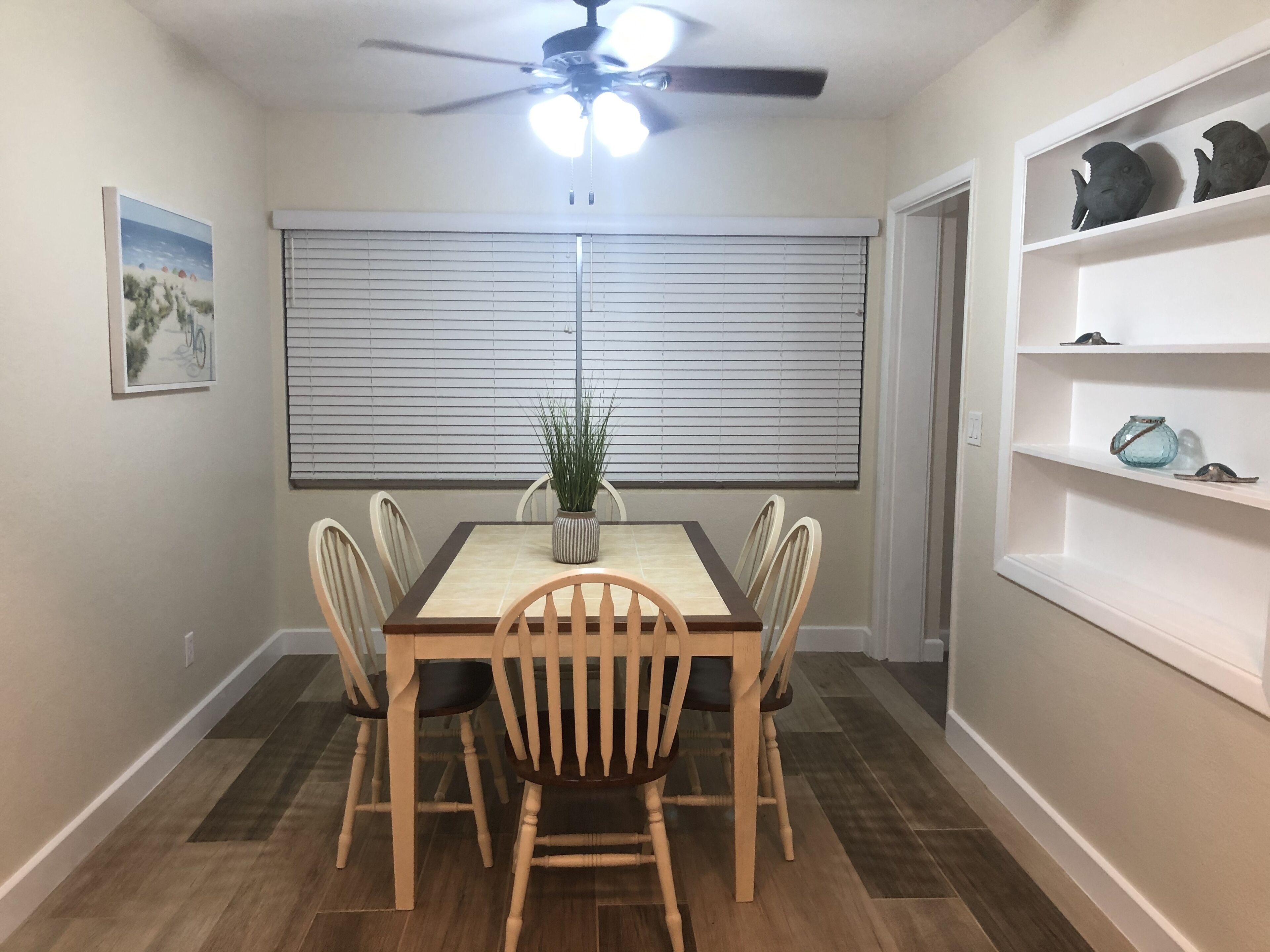 A dining area with a table, chairs, a ceiling fan, a window with blinds, and a shelf with decorative items.