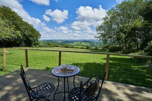 A deck with a table and chairs overlooking a grassy field and hills. - Pencastell Views Pod With Stunning Views (Lampeter)