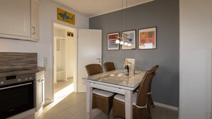 A modern kitchen with white cabinets, a built-in oven, and a dining area with wicker chairs and a glass table. - Haus Möwe apartment 5/13 (Hörnum (Sylt))