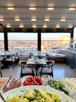 Un restaurant avec des tables et des chaises, un buffet proposant divers plats, et une vue sur la ville grâce à de grandes fenêtres.