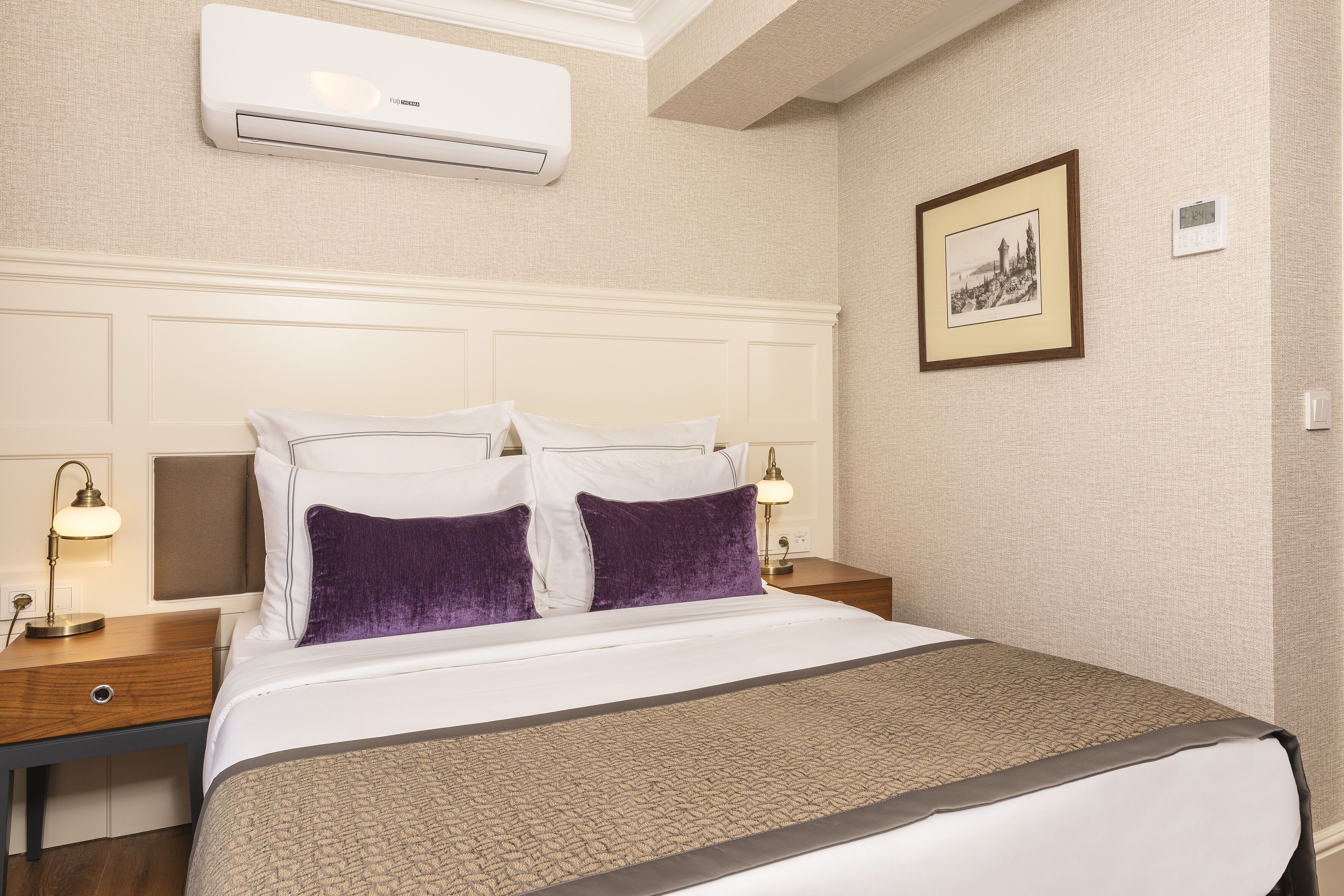 A hotel room with a large bed, two bedside tables with lamps, a wall-mounted air conditioner, a framed picture, and a thermostat.