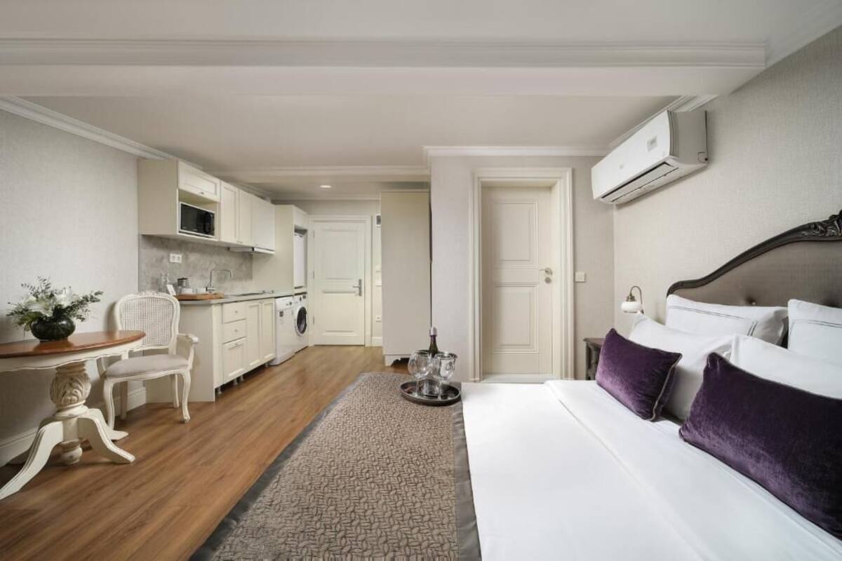 A hotel room with a bed, a dining area with a table and chairs, a kitchen with cabinets and a microwave, and an air conditioning unit on the wall.